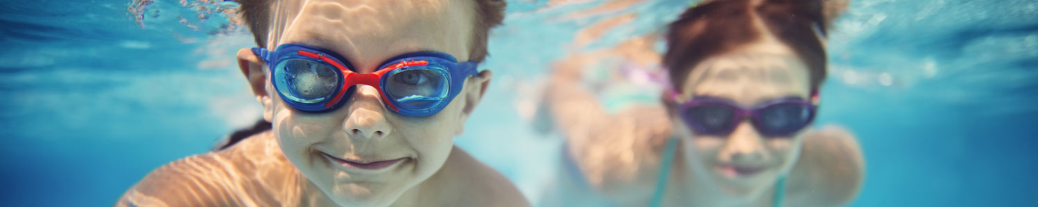 A boy in goggles smiles underwater with a girl swimming behind him.