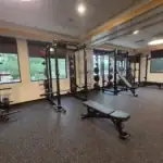 A gym interior featuring squat racks, weight plates, a bench, and large windows.
