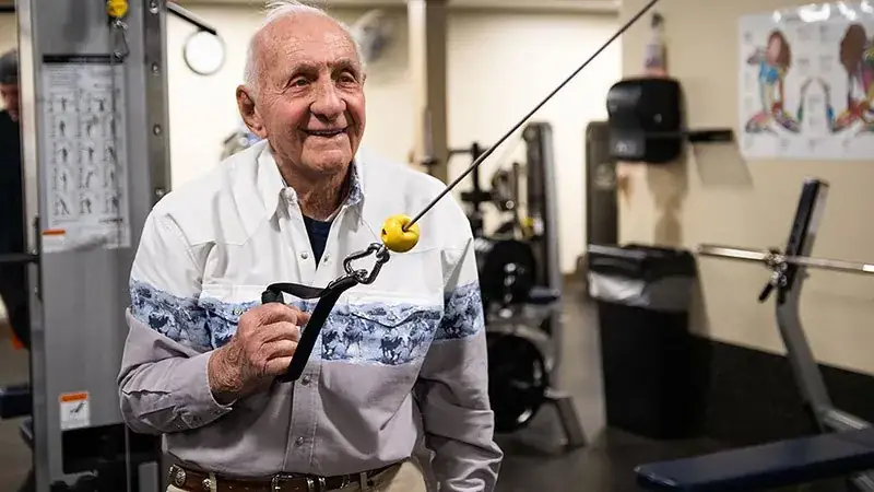 An elderly man exercises with a cable machine in a gym, wearing a patterned shirt and smiling.
