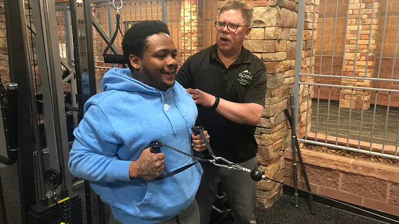 A person in a blue hoodie exercises on a cable machine, assisted by another person in glasses and a black shirt.