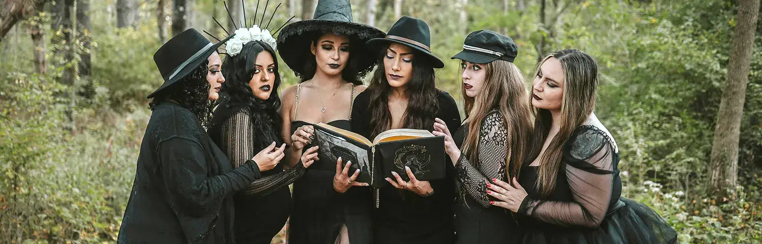A group of six individuals dressed in black, gather in a forest, examining a large open book.