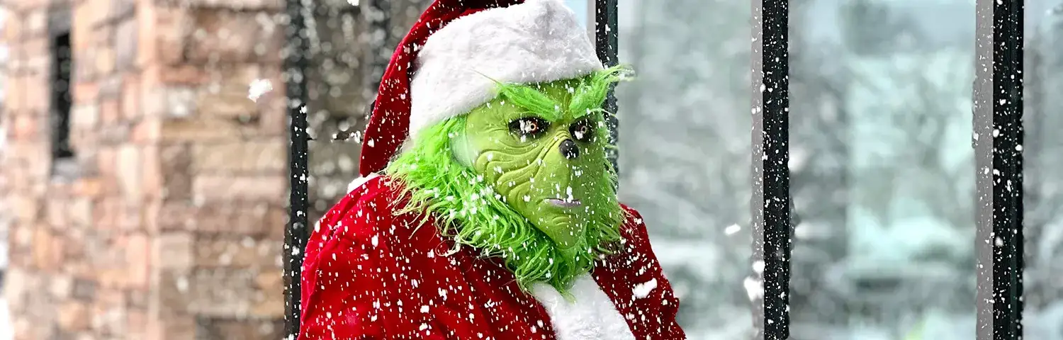 Person dressed as the Grinch in a Santa outfit stands outside a building, holding out hands while snow falls around.
