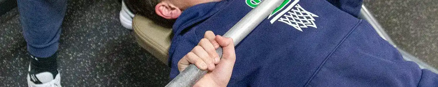 A young person is lying on a bench press bench in a gym, gripping a barbell. Another person stands nearby.