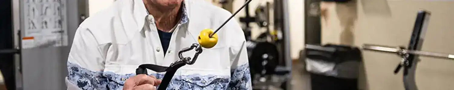 An elderly man exercises with a cable machine in a gym, wearing a patterned shirt and smiling.