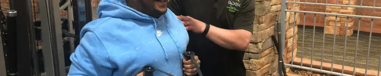 A person in a blue hoodie exercises on a cable machine, assisted by another person in glasses and a black shirt.