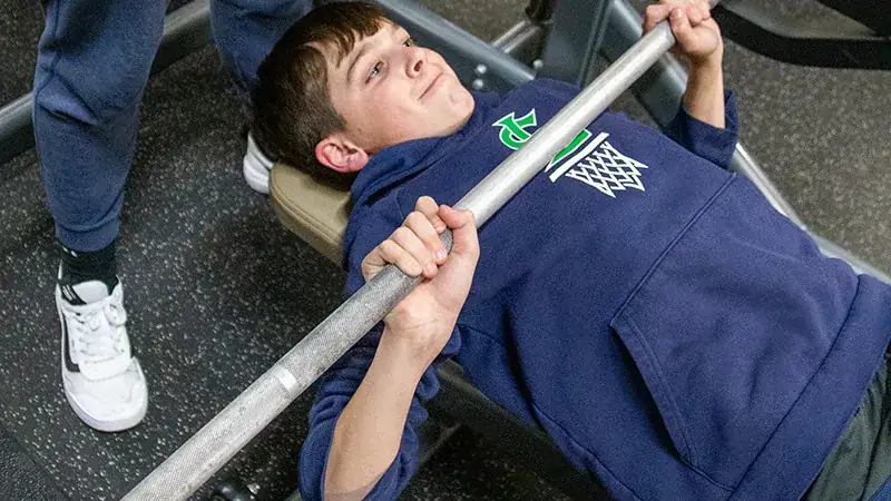 A young person is lying on a bench press bench in a gym, gripping a barbell. Another person stands nearby.
