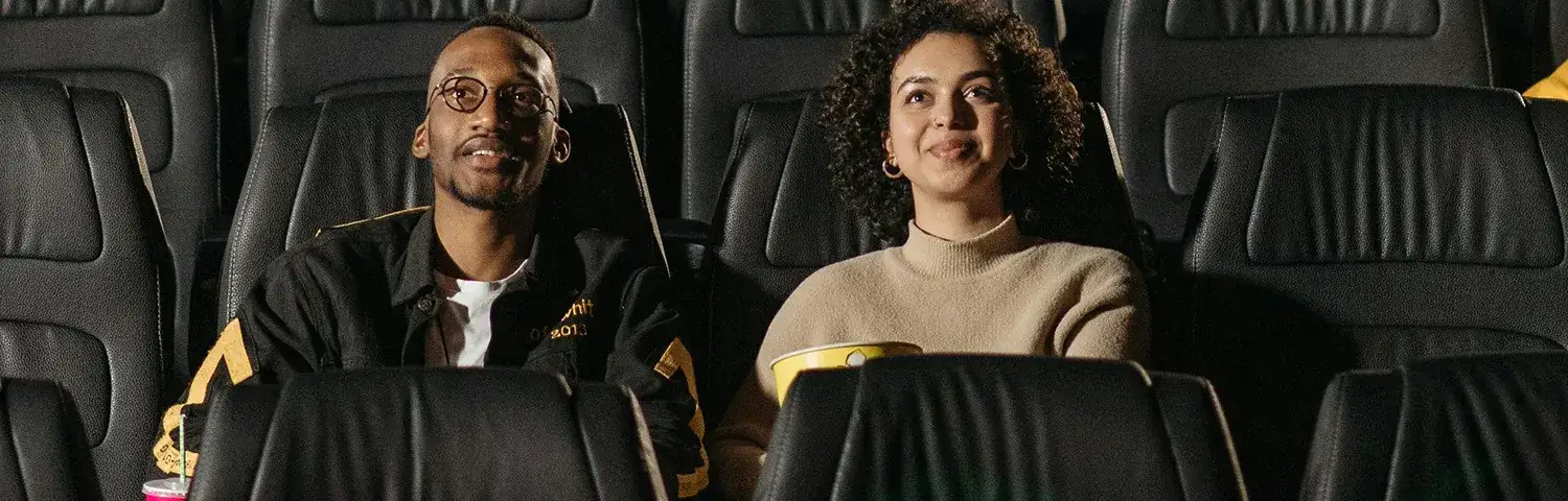 Two people seated in plush cinema chairs, enjoying a movie with popcorn and drinks.