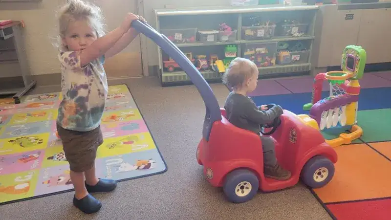 A toddler pushes a younger child in a red ride-on toy car.