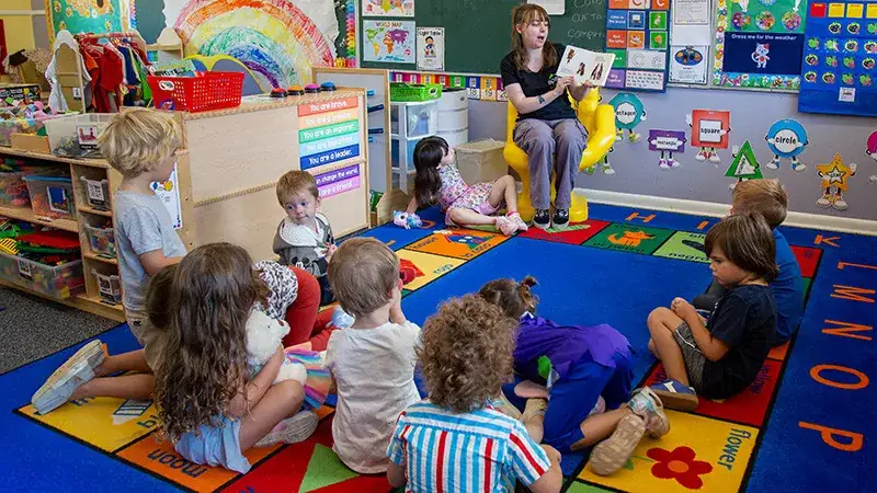 A teacher reads to a group of young children sitting on a colorful rug.