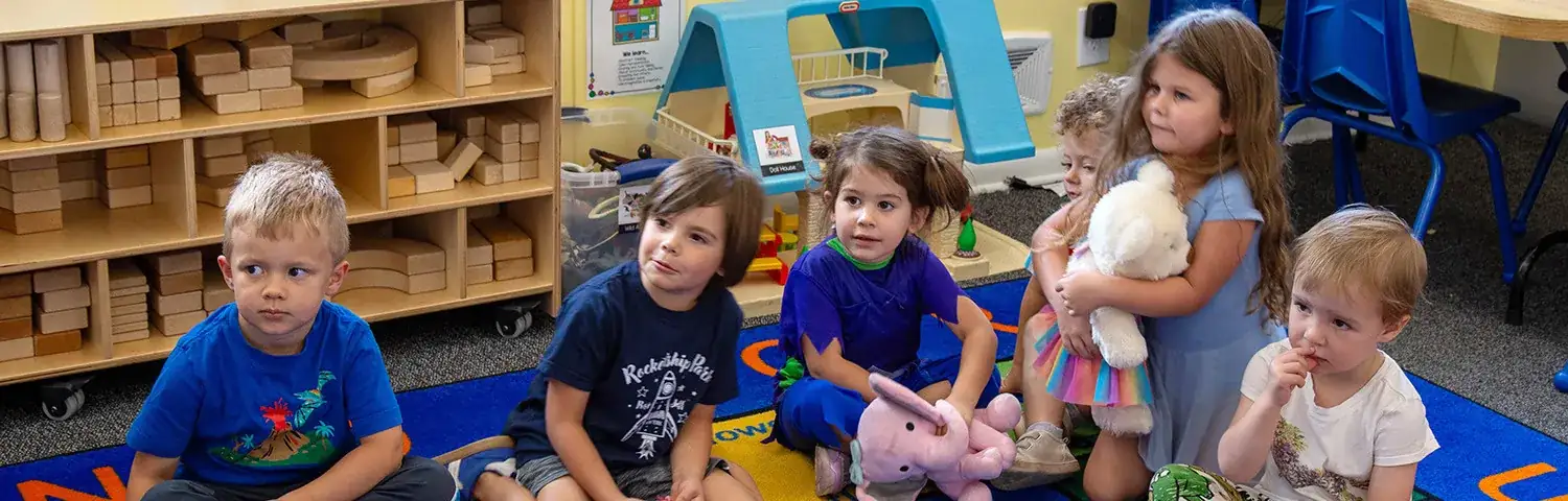 A group of children sit on a colorful carpet in a classroom.
