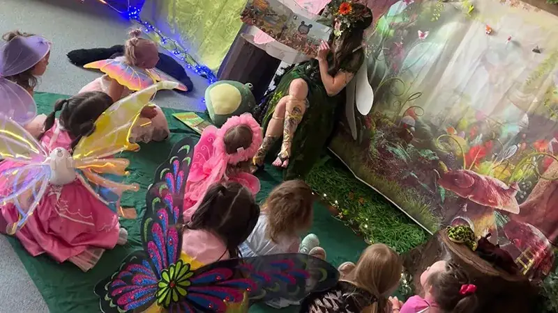 Children in fairy costumes sit on a green carpet, led by a woman dressed as a fairy.