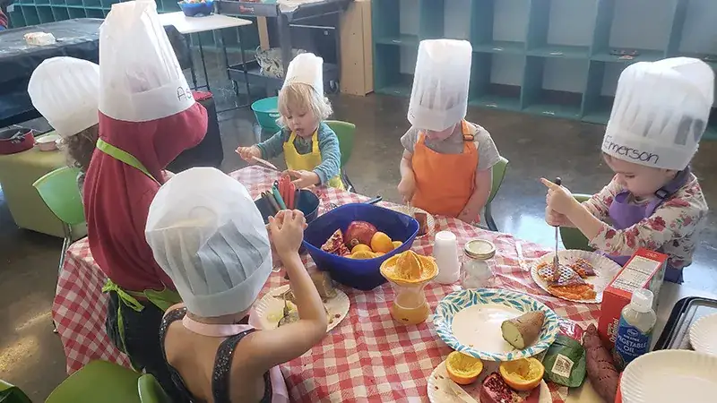 Children in chef hats are gathered around a table, engaged in a cooking activity.