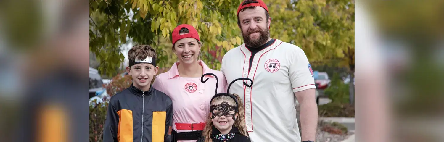 Four people stand outdoors, dressed in costumes, surrounded by fall foliage.