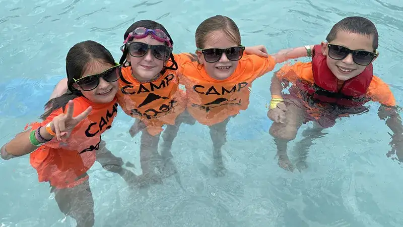 Four kids in bright orange "CAMP" shirts play in shallow water.