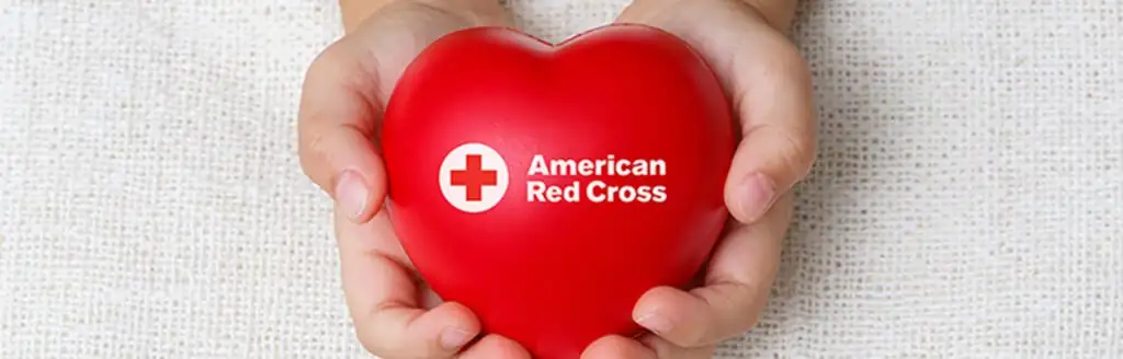 Hands holding a red heart-shaped object with the American Red Cross logo.