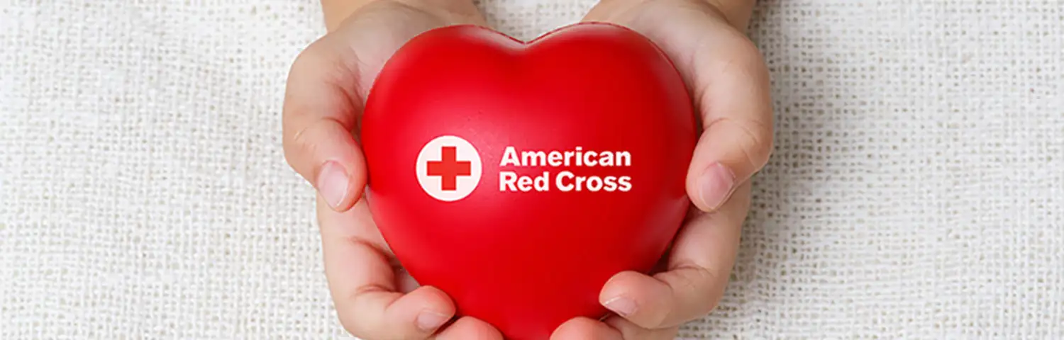 Hands holding a red heart-shaped object with the American Red Cross logo.