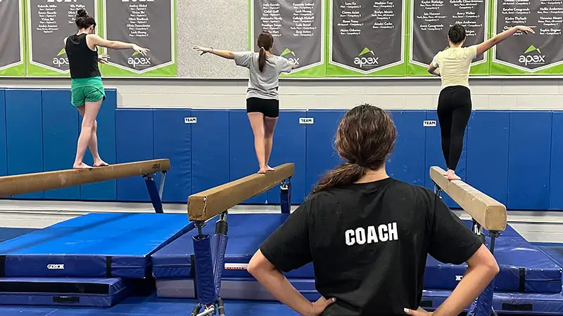 Coach watches three gymnasts balancing on separate uneven bars in a gym with gymnastics state champion banners displayed on the wall.