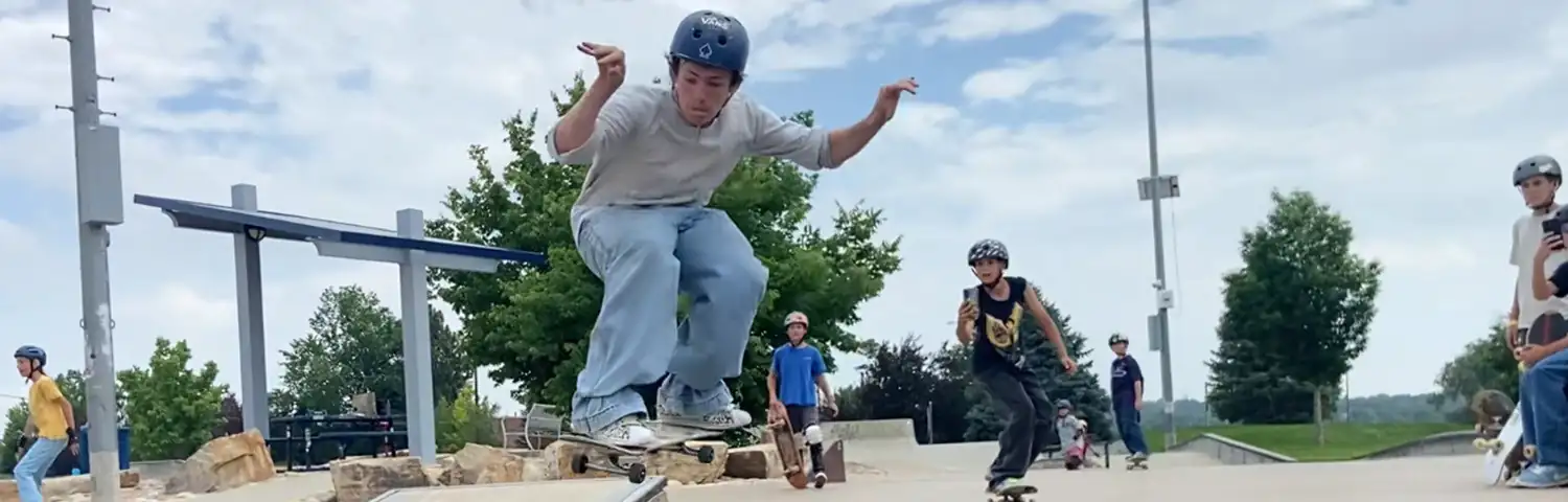 Teen skateboarder in grey helmet and baggy clothes performs trick at outdoor skate park with other skaters watching.