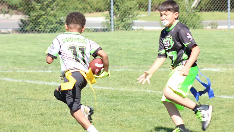Youth flag football player carrying the ball while another player runs toward them on a grass field.
