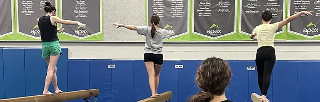 Coach watches three gymnasts balancing on separate uneven bars in a gym with gymnastics state champion banners displayed on the wall.