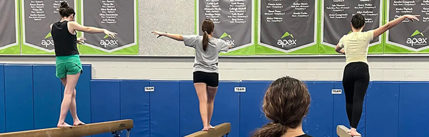 Coach watches three gymnasts balancing on separate uneven bars in a gym with gymnastics state champion banners displayed on the wall.