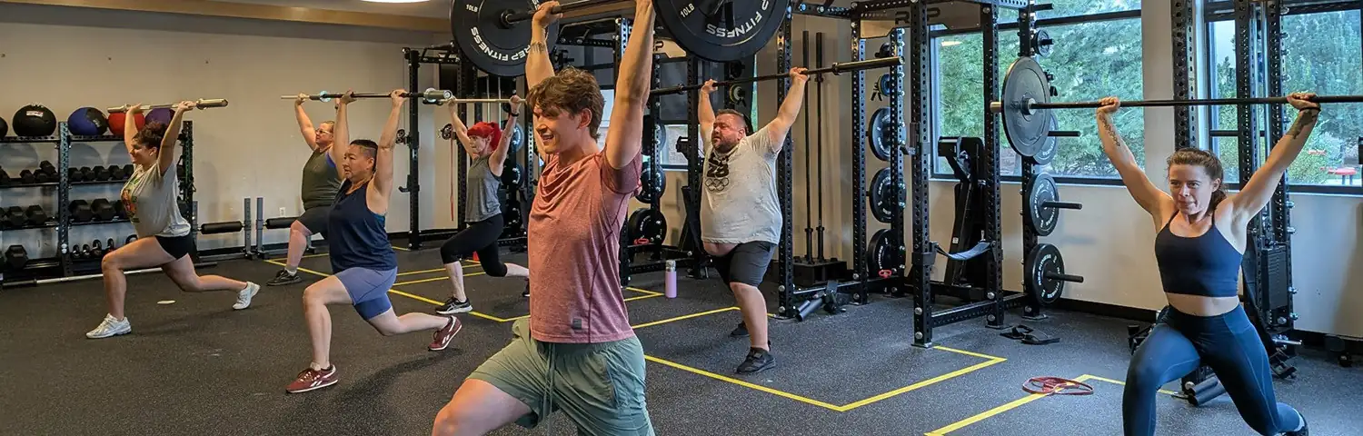 Group of people performing lunges in a gym with weight racks and large windows.