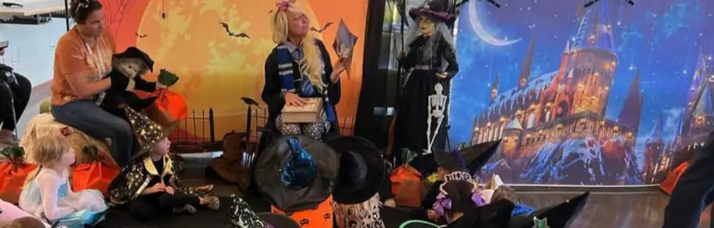 Children and adults in Halloween costumes pose in a decorated room with witches, pumpkins, bats, and a castle backdrop.