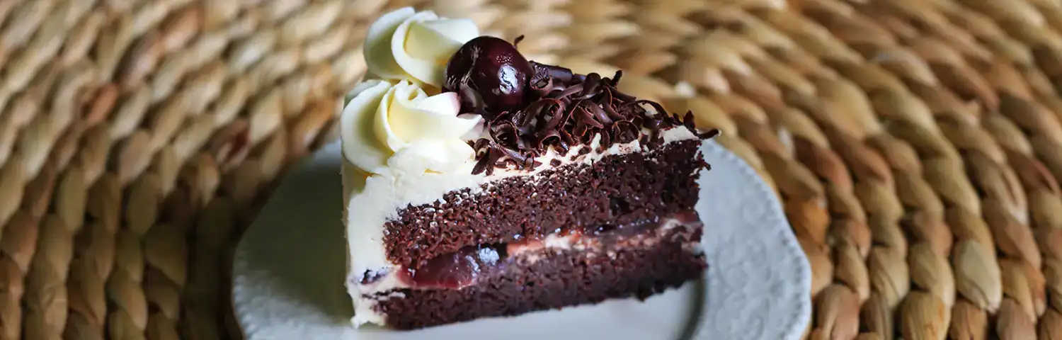 Slice of chocolate cake with whipped cream, cherries, and chocolate shavings on a white patterned plate.