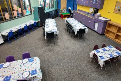 Classroom with tables covered in party-themed tablecloths and plates, surrounded by colorful chairs, cabinets, and children's artwork.