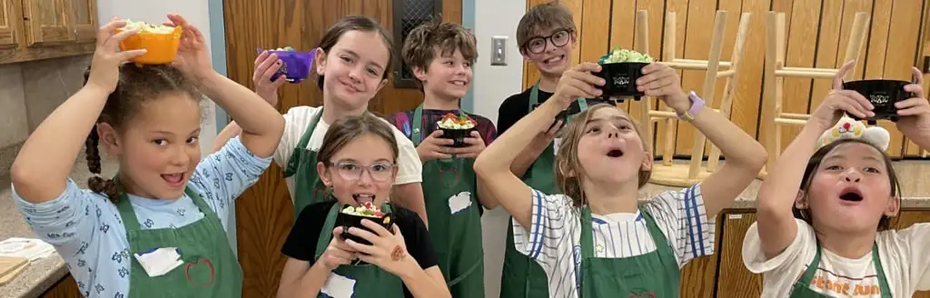 Seven children in aprons joyfully hold up bowls of food during a cooking activity.