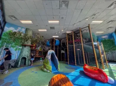 Indoor children's play area with colorful slides, climbing structures, and tree-themed decor, featuring artificial grass flooring and kids playing.