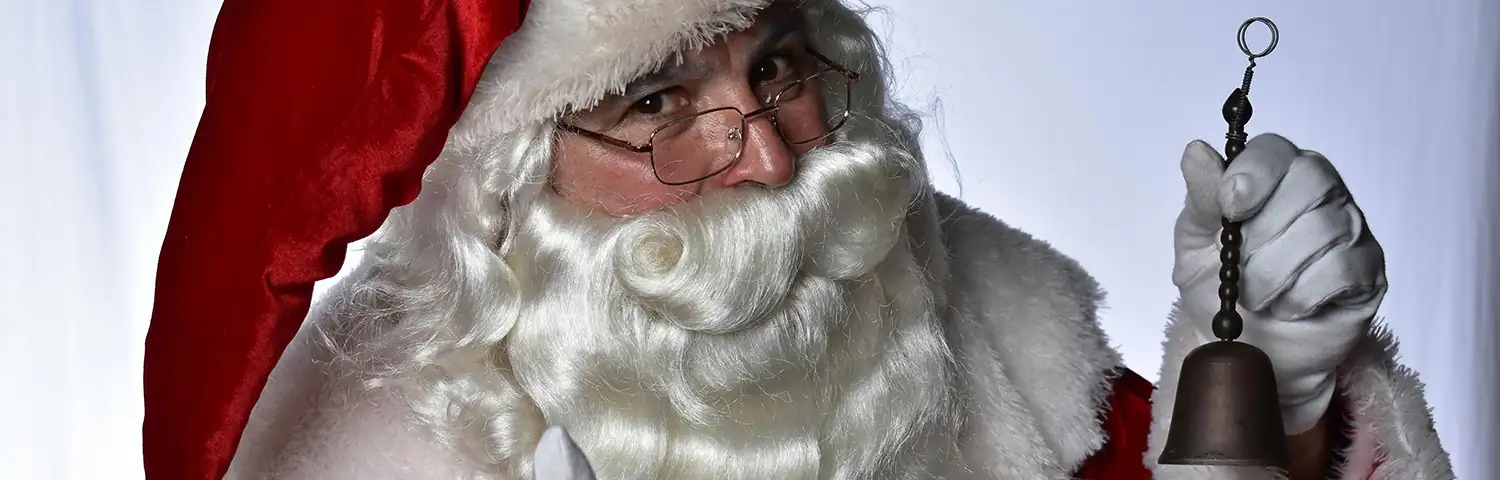 Close-up of Santa Claus wearing glasses, a red hat, and white gloves, holding a small metal bell.