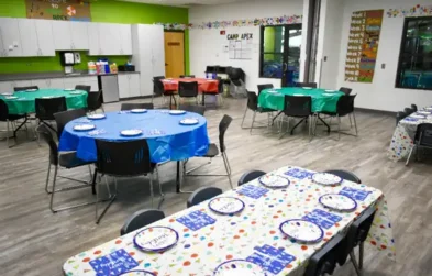 Classroom set up for a party with tables covered in colorful tablecloths, plates, and napkins, with chairs arranged around them.