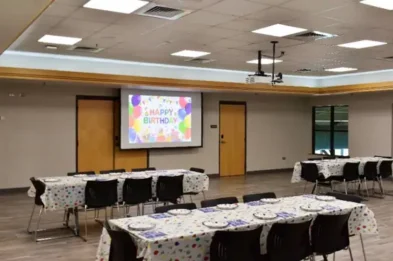 Birthday party setup in a large room with decorated tables, chairs, and a projector displaying a colorful Happy Birthday message.