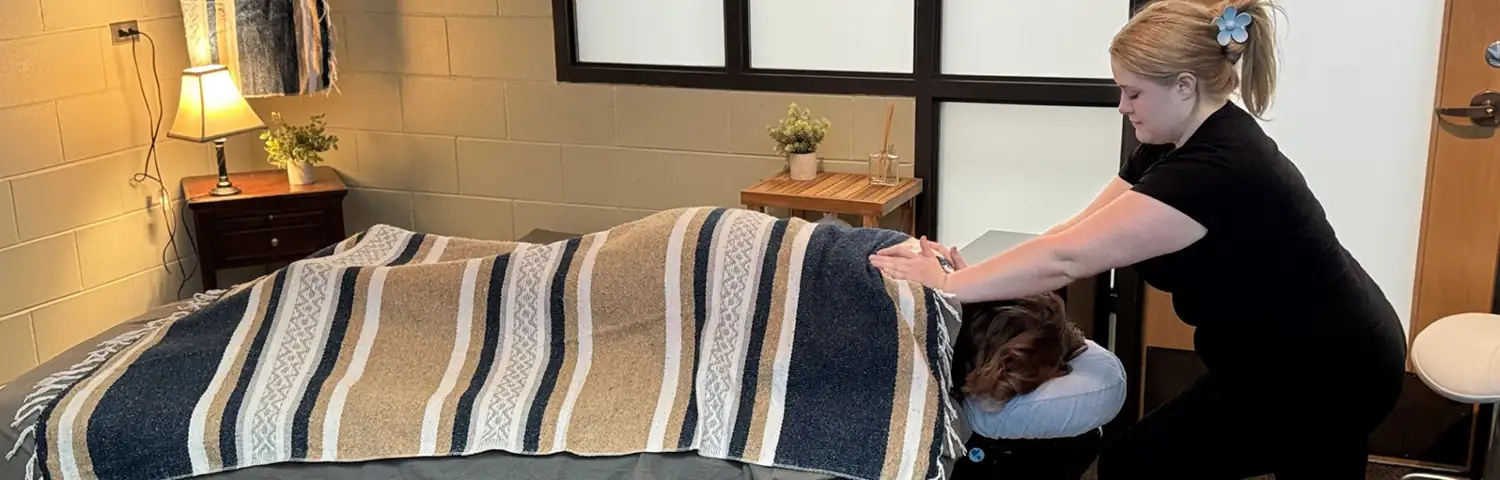 A woman receives a back massage while lying face down on a massage table, covered with a striped blanket.
