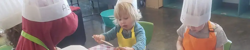 Children in chef hats are gathered around a table, engaged in a cooking activity.