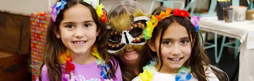 Two girls and an adult in colorful flower leis and festive masks.