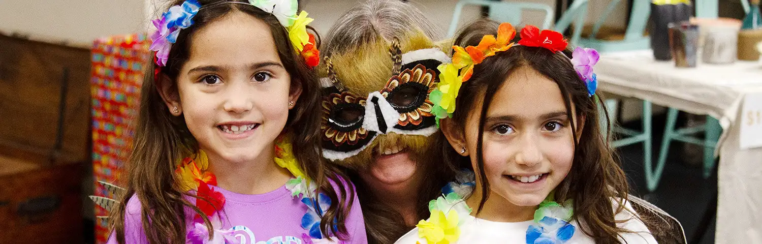 Two girls and an adult in colorful flower leis and festive masks.