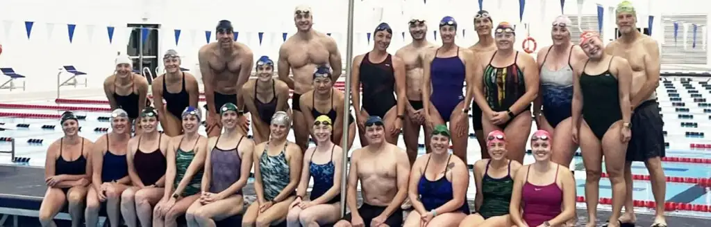 Group of swimmers in swimsuits and swim caps posing beside an indoor pool.