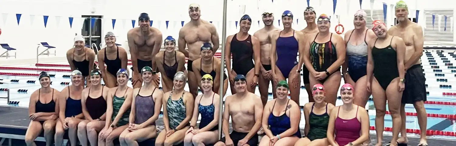 Group of swimmers in swimsuits and swim caps posing beside an indoor pool.