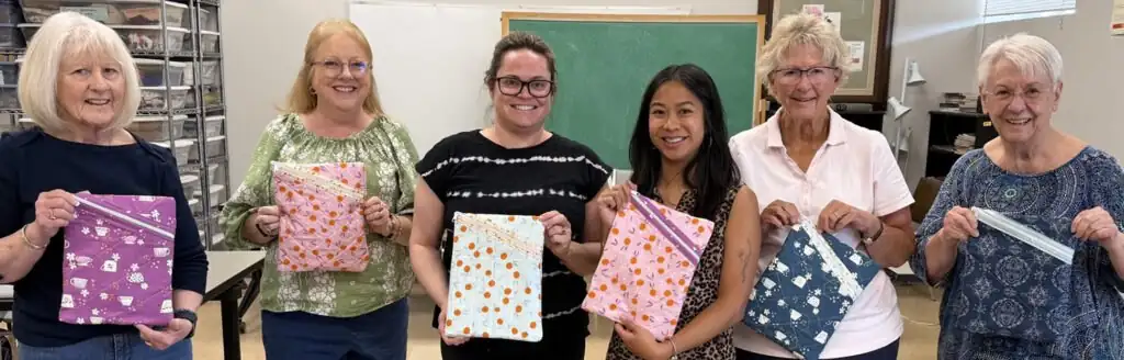 Six women smile while holding handmade zippered fabric pouches in a craft room.